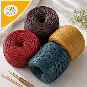 May include: Four round balls of raffia yarn in dark brown, deep red, mustard yellow, and teal blue, arranged together. Two metal crochet hooks with wooden handles are positioned at the bottom. The logo CHILL FUZZ is visible in the top corner.