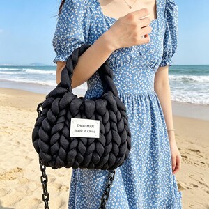Op de afbeelding: Een zwarte, dikgebreide handtas met een kettingriem. De tas heeft een gevlochten handvat en een rechthoekig label met de tekst "ZHOU MAN Made in China". De achtergrond is een strand en de oceaan.
