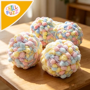 Peut inclure: Trois pelotes de laine colorées avec des pompons aux tons pastel de rose, jaune, bleu et vert reposent sur une surface en bois. La laine est duveteuse et texturée, idéale pour les projets de tricot ou de crochet. L'image comprend le texte "CHILL FUZZ".