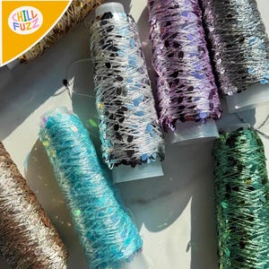 Puede incluir: Primer plano de varios carretes de hilo decorativo en varios colores, incluyendo plata, turquesa, morado y verde. Cada carrete está envuelto con lentejuelas brillantes, creando un efecto texturizado y llamativo. El logo "CHILL FUZZ" es visible en la esquina superior izquierda.