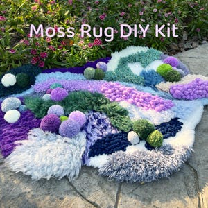 May include: A handmade moss rug DIY kit with a variety of textures and colors. The rug features shades of purple, green, blue, and white, with pom-poms and textured yarn. The text "Moss Rug DIY Kit" is displayed at the top.
