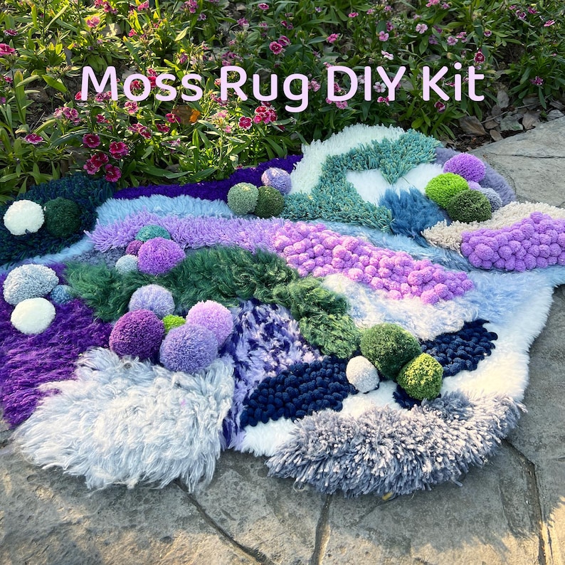 May include: A handmade moss rug DIY kit with a variety of textures and colors. The rug features shades of purple, green, blue, and white, with pom-poms and textured yarn. The text "Moss Rug DIY Kit" is displayed at the top.