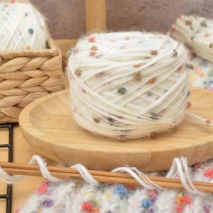 May include: A close-up of white yarn with colorful flecks, resting on a wooden plate. The yarn is in a ball shape, with a basket of yarn in the background. Knitting needles and a knitted piece are in the foreground.