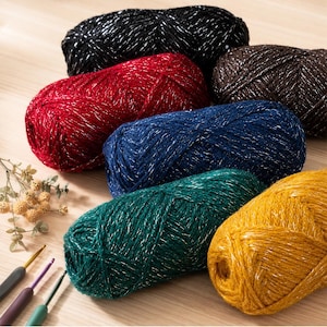 May include: Six skeins of yarn in black, red, brown, dark blue, dark green, and mustard yellow, each featuring a metallic silver thread woven throughout. Three crochet hooks with colored handles are positioned near the yarn skeins.