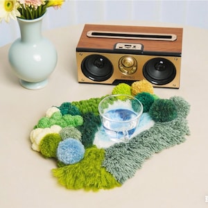 May include: A handmade, colorful coaster with a glass of water, featuring a mix of green, blue, and yellow pom-poms and fringe. A wooden and gold speaker sits in the background, along with a vase of flowers.