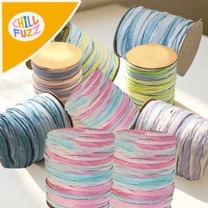 Pode incluir: Uma coleção de rolos de fita de ráfia colorida em vários tons pastel. Os rolos estão dispostos em grupo, mostrando as diferentes combinações de cores, incluindo azul, rosa e verde. O logotipo "CHILL FUZZ" é visível no canto superior esquerdo.