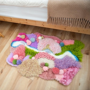 K&ouml;nnte beinhalten: Ein farbenfroher, handgefertigter Teppich mit einem skurrilen Design. Der Teppich weist verschiedene Texturen und Farben auf, darunter Rosa, Lila, Gr&uuml;n und Wei&szlig;, mit Bommelakzenten. Der Teppich liegt auf einem Holzboden in der N&auml;he eines Bettes.