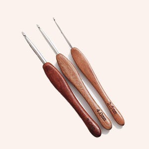 May include: Three crochet hooks with wooden handles in varying shades of brown. The metal hook ends are silver. The handles are labeled with sizes: 5.0mm, 4.0mm, and 2.5mm. The hooks are arranged diagonally against a light background.