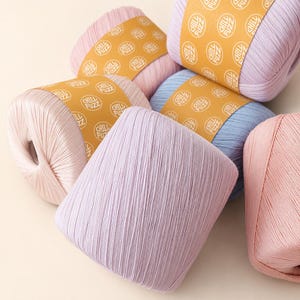 May include: Five skeins of yarn in pastel shades of light pink, lavender, and baby blue. Each skein is wrapped in a mustard yellow paper band featuring the repeating white text Chill Fuzz. The yarn has a smooth, tightly wound texture.