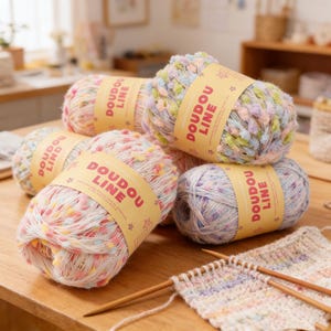 Puede incluir: Varias madejas de lana colorida, cada una con la etiqueta "DOU DOU LINE". La lana es una mezcla de tonos pastel, incluyendo rosa, amarillo, azul y verde. También se ven agujas de tejer y una muestra parcialmente tejida.