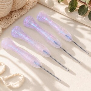 May include: Four crochet hooks with iridescent, glittery lavender handles and silver metal hooks. The handles have a textured, pearlescent appearance. The hooks are arranged diagonally, showcasing their design and craftsmanship. Ideal for crochet enthusiasts.