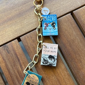 May include: A gold-colored keychain with three miniature book charms. The books are titled "Heated Rivalry", "Call Me By Your Name", and "The Song of Achilles". The keychain also has a gold clasp and a small white charm.