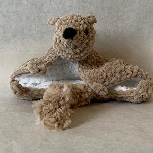 May include: A brown crocheted stuffed animal with a white fleece lining. The animal has a small tail and a face with black eyes and a black nose.
