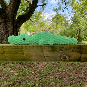 Puede incluir: Un juguete de peluche de cocodrilo verde tejido a crochet que se encuentra sobre una valla de madera. El cocodrilo tiene ojos negros y una superficie texturizada.