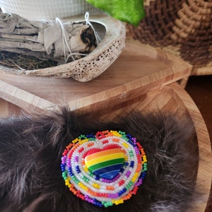 May include: A heart-shaped beaded pin with a rainbow design in the center, surrounded by layers of white, orange, yellow, green, blue, and purple beads. The pin is set on a bed of dark brown feathers, resting in a wooden bowl.