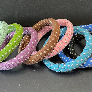 May include: A collection of colorful bangles in shades of green, purple, brown, pink, blue, and black. Each bangle is wrapped in fabric and adorned with evenly spaced silver-colored studs. The bangles are stacked together, showcasing their variety.