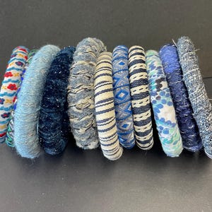May include: A collection of fabric-covered bracelets in various shades of blue, white, and red. The bracelets feature diverse patterns, including floral, geometric, and textured designs. The assortment showcases a range of styles, from solid colors to intricate woven details.