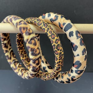 May include: Three leopard print bangles. The bangles are in varying shades of tan, brown, and black. The bangles are made of different materials, including a soft, plush fabric and a ribbon-like material. The bangles are displayed on a wooden dowel.
