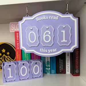 Peut inclure: Un compteur de livres de couleur lavande avec le texte "Books read this year" et les chiffres "061". Un compteur séparé affiche "100". Les compteurs sont affichés sur une étagère avec des livres.