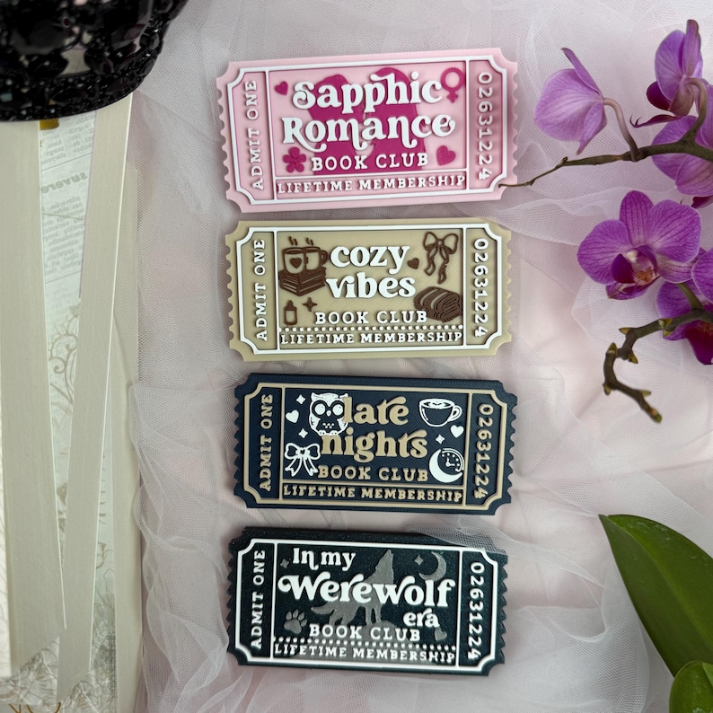 K&ouml;nnte beinhalten: Vier dekorative Magnete in Ticketform mit den Texten "Sapphic Romance Book Club", "Cozy Vibes Book Club", "Late Nights Book Club" und "In my Werewolf Era Book Club". Jeder Magnet hat ein einzigartiges Design und Farbschema, darunter Rosa, Beige, Marineblau und Schwarz.