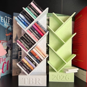 Può includere: Due librerie decorative, una bianca e una verde chiaro, ciascuna con un design unico a forma di albero. La libreria bianca contiene diversi libri, mentre la libreria verde è vuota. La libreria bianca ha "TBR" inciso sulla base.