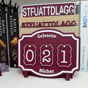 Peut inclure: Un compte-livres bordeaux et blanc affichant le nombre "021". Les mots "Gelesene" et "Bücher" sont affichés au-dessus et en dessous des chiffres. Le compteur est sur une étagère avec des livres et des enseignes décoratives.