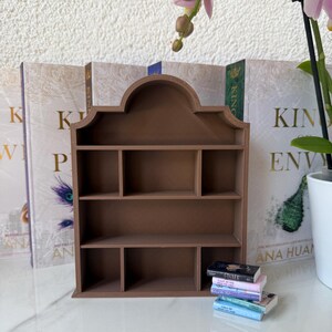 Può includere: Una piccola libreria in legno marrone con diversi scomparti. Una pila di libri in miniatura è appoggiata alla base. Lo sfondo presenta copertine di libri e un vaso bianco con una pianta.