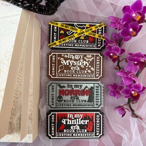 K&ouml;nnte beinhalten: Vier magnetische Tickets mit Text: "Crime Scene", "Mystery era", "Horror era" und "Thriller era" Buchclub. Die Magnete sind schwarz, braun und rot mit wei&szlig;em Text. Violette Orchideen sind im Hintergrund.