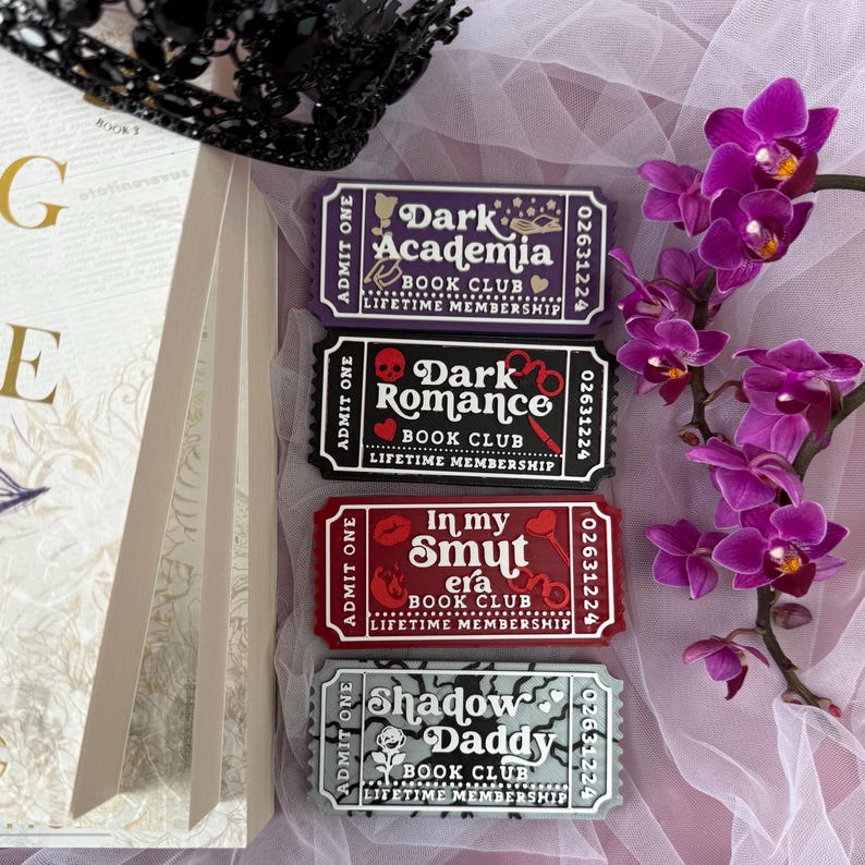 K&ouml;nnte beinhalten: Vier rechteckige Lesezeichen mit Ticket-Designs. Die Lesezeichen tragen die Aufschriften "Dark Academia", "Dark Romance", "In my Smut era" und "Shadow Daddy". Jedes Lesezeichen hat ein einzigartiges Farbschema und Design. Ein Zweig lila Orchideen und ein Buch sind ebenfalls im Bild.
