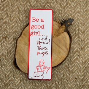 Può includere: Segnalibro rosso con il testo "Be a good girl... and spread those pages." e un disegno a linea di una persona che legge un libro.