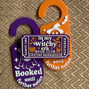 Könnte beinhalten: Zwei Türhänger mit Hexen-Motiv. Einer ist lila mit dem Text "Booked until further notice". Der andere ist orange mit einem Hexenhut und Fledermäusen. Ein Ticket-förmiges Etikett lautet "In my Witchy era BOOK CLUB LIFETIME MEMBERSHIP."
