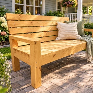Puede incluir: Un banco de madera de tono claro natural con respaldo de listones y asiento plano con patas cuadradas robustas. El banco está decorado con un cojín cuadrado de color crema con textura, un cojín verde oliva liso y una manta verde con patrón de espiga.