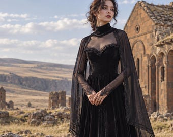 May include: A floor-length black velvet dress featuring a sweetheart neckline, a high mock neck collar, and long sheer mesh sleeves. The garment includes a matching sheer mesh cape overlay with delicate lace trim along the edges and shoulders.