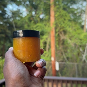 May include: A small, clear jar with a black lid filled with a bright orange, thick, gel-like substance. The jar is being held in a person's hand.