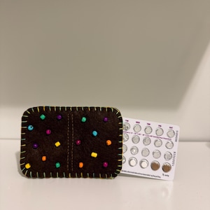 May include: A brown felt wallet decorated with colorful beads and a yellow stitched border. Next to the wallet is a blister pack of birth control pills. The wallet is rectangular and has a vertical seam down the middle.