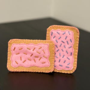 May include: Two felt food items resembling frosted pastries. The pastries have a pink frosting with bead sprinkles and a tan crust. One is rectangular, and the other is more square. The edges are stitched with a tan thread.