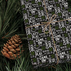 May include: A black gift box wrapped in paper with a repeating pattern of white text that says "Game On", "Level Up", "Lets Play", and "Game Over" with green accents. The box is tied with twine.