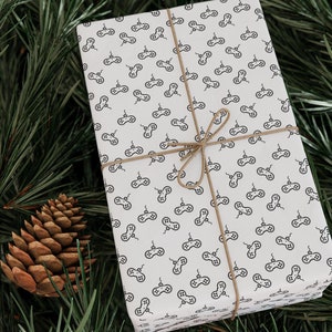 May include: A white gift box with a brown twine bow. The box is wrapped in paper with a repeating pattern of black video game controllers.