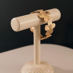 Peut inclure: Un bracelet floral doré présenté sur un présentoir à bijoux crème. Le bracelet est composé de plusieurs maillons en forme de fleurs. Le présentoir a un dessus cylindrique et une base décorative avec un motif tourbillonnant. Le fond est de couleur neutre foncée.