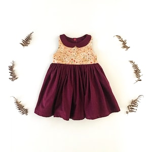 May include: A sleeveless dress with a burgundy skirt and a floral bodice. The bodice has a rounded collar and a button closure at the back. The dress is surrounded by dried ferns on a white background.