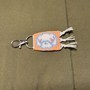 May include: A small, orange and white woven keychain with a white cloud design and blue eyes. The keychain has a silver metal ring and white tassels.