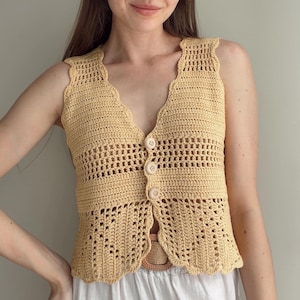 May include: A pale yellow sleeveless crochet vest featuring a V-neckline, a scalloped edge, and three light-colored circular buttons down the center. The garment is crafted with a combination of solid stitches and an openwork lace pattern.