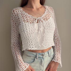 Può includere: Un bolero all'uncinetto bianco a maniche lunghe con un motivo a rete aperta e chiusura frontale a laccio. Il capo è corto, presenta maniche svasate ed è indossato sopra una canotta bianca semplice abbinata a jeans in denim azzurro.