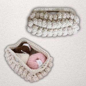 May include: Two cream-coloured, handmade crochet baskets. One is filled with brown, white, and pink yarn. The other is empty. The baskets feature a textured, bubble-like design and are suitable for storing craft supplies.
