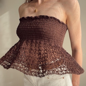 Puede incluir: Un top de crochet sin tirantes de color marrón oscuro que presenta un corpiño ajustado y acanalado con un borde superior con volantes y un dobladillo de peplum de encaje calado. La prenda está confeccionada con un punto denso y un diseño aireado.