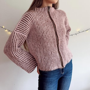 May include: A dusty pink, long-sleeved, ribbed knit jumper featuring a high mock neck collar and a full-length front zip fastening. The garment has a textured, vertical ribbed pattern throughout the body and sleeves, with a straight hemline.