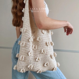 Pode incluir: Uma bolsa tote de crochê bege com um design de flores. A bolsa tem uma parte superior grande e aberta e uma única alça. A bolsa é feita à mão com amor, como indicado pelo texto "ALYA LOOM Handmade with love" na bolsa.