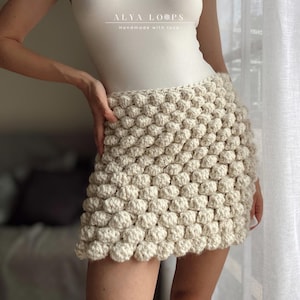 May include: Cream-coloured crochet mini skirt with a textured, bubble-like pattern. The skirt is handmade and has a fitted design. The text "ALYA LOOPS Handmade with love" is at the top.