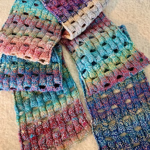 May include: A hand-knitted scarf featuring a basketweave pattern with an open-work design. The yarn displays a variegated color palette of blue, purple, teal, and red tones, with a solid light gray section at one end of the rectangular accessory.