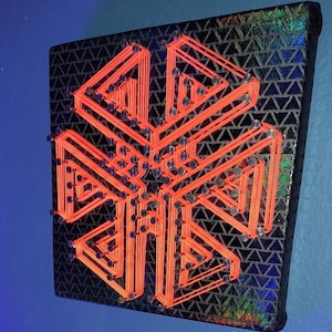 May include: A geometric string art piece on a black canvas. The design features a symmetrical pattern in bright orange thread, secured with small silver nails. The background has a textured, iridescent pattern.